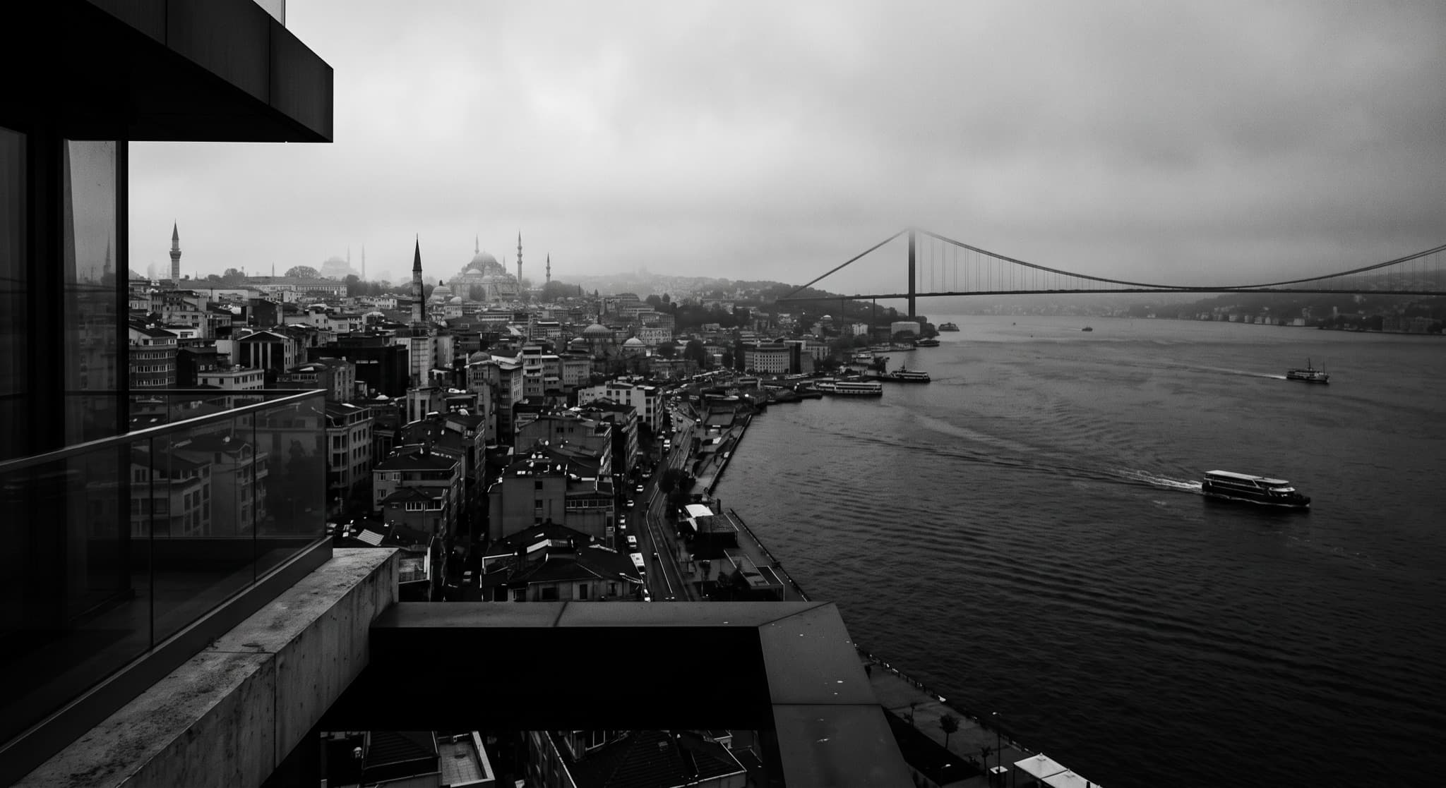 Black-and-white view over the Golden Horn from a concrete terrace, the Istanbul skyline and a suspension bridge fading into mist.